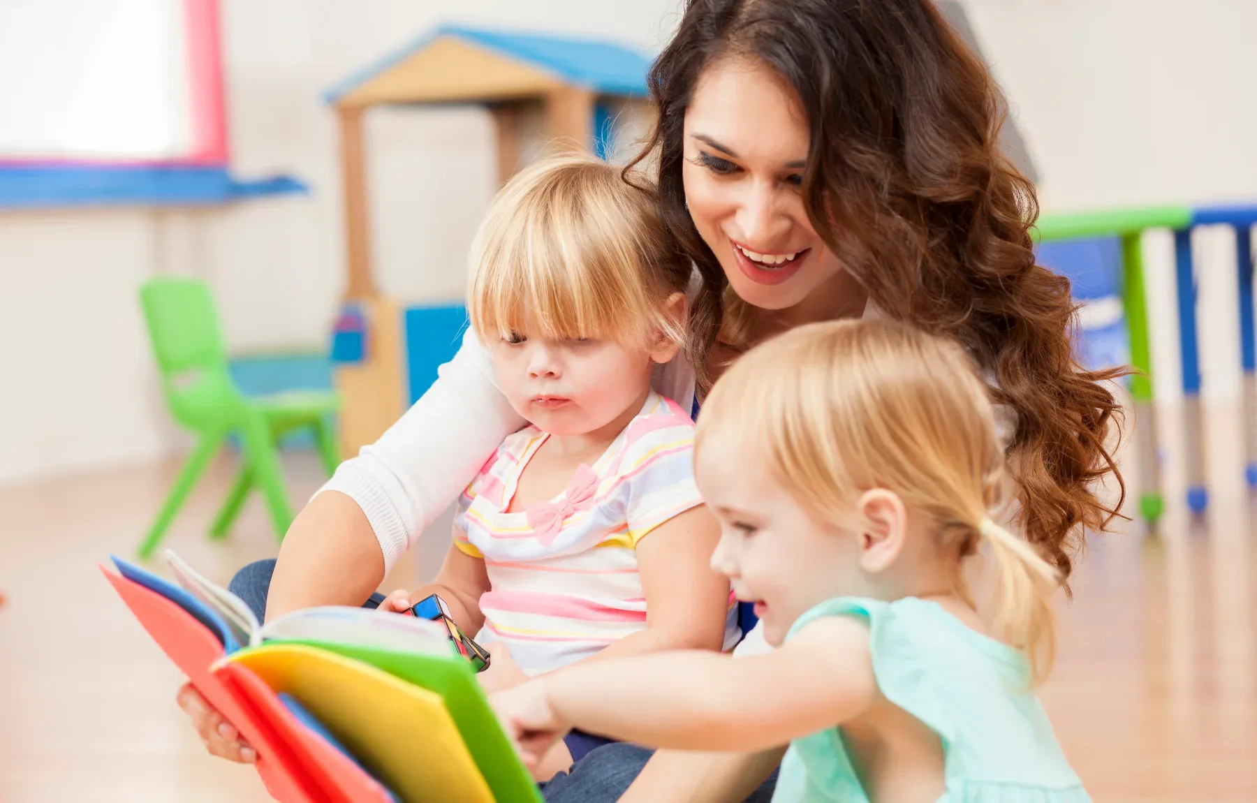 Smiling caregiver reading a colorful book with two toddlers in a bright, playful daycare classroom environment.
